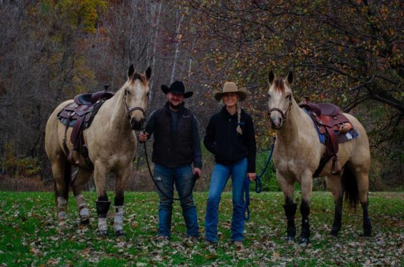 Two riders stand beside their horses in a forested area during autumn.